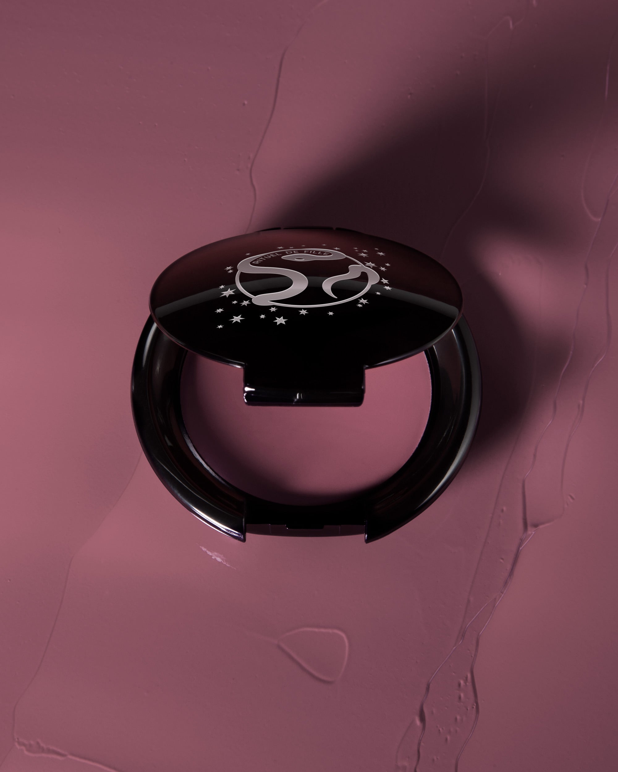 A round black makeup compact with a glossy lid featuring a white stylized logo and stars, set against a textured mauve-pink background with a matching makeup smear.