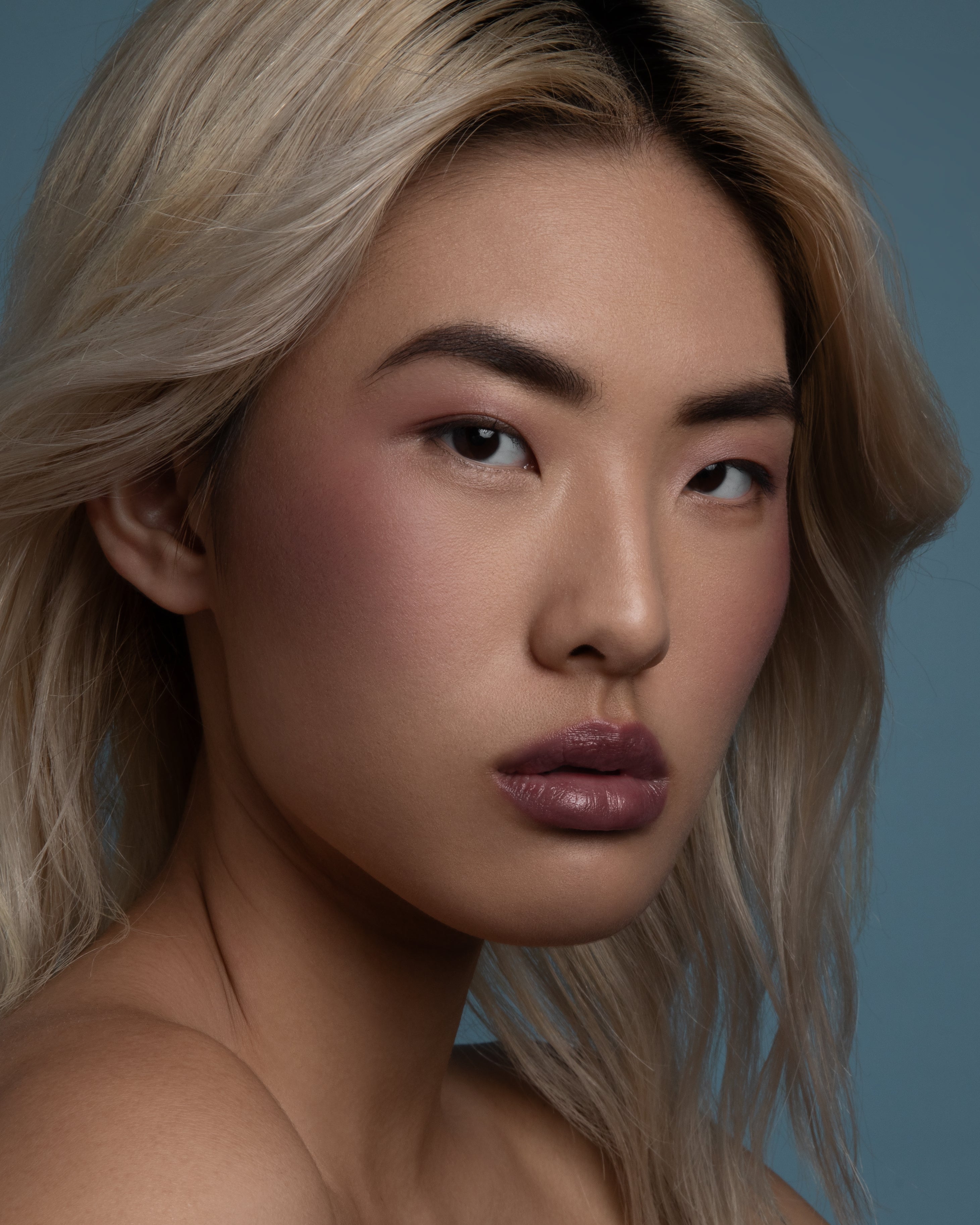 A close-up portrait of a woman with blonde hair, wearing a glossy mauve-pink lip color, set against a solid slate blue background.