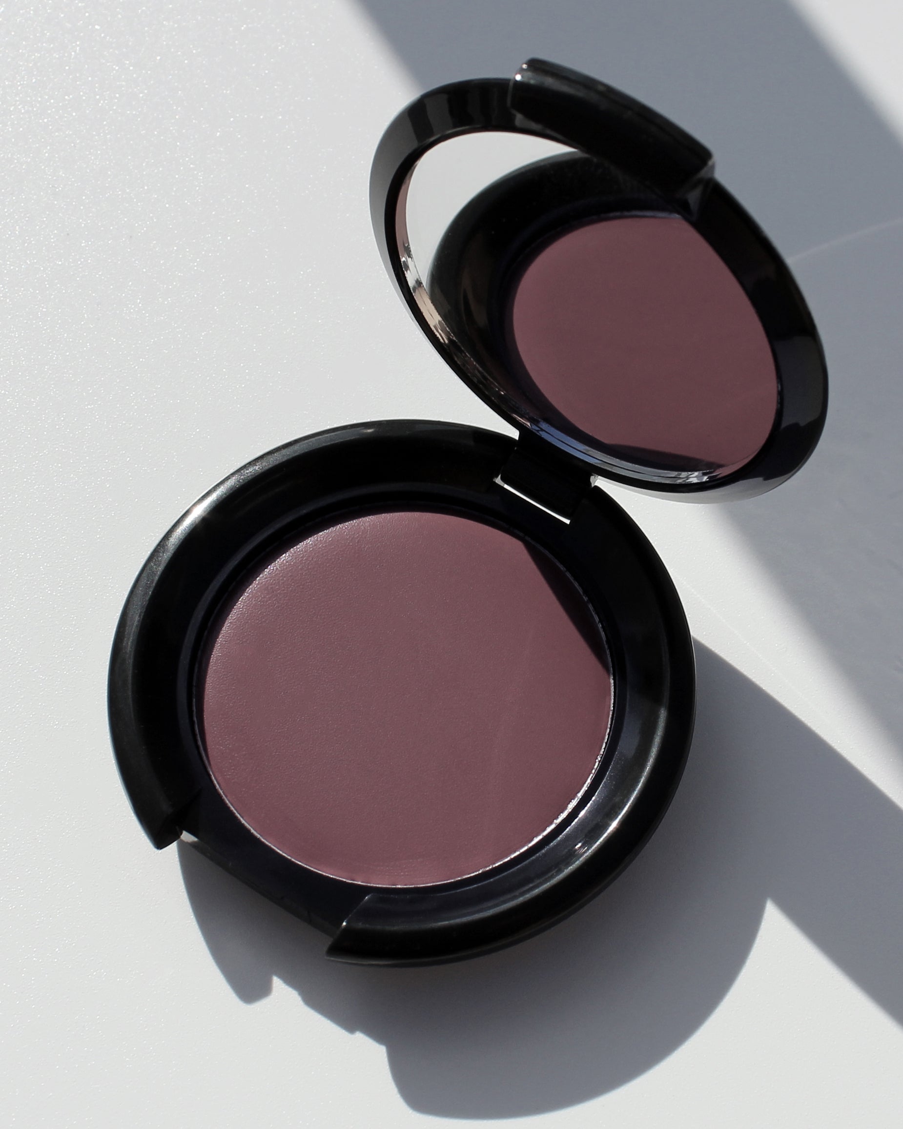 An open round makeup compact with a black glossy case and a mauve-pink cream blush, photographed on a light gray surface with strong shadows.